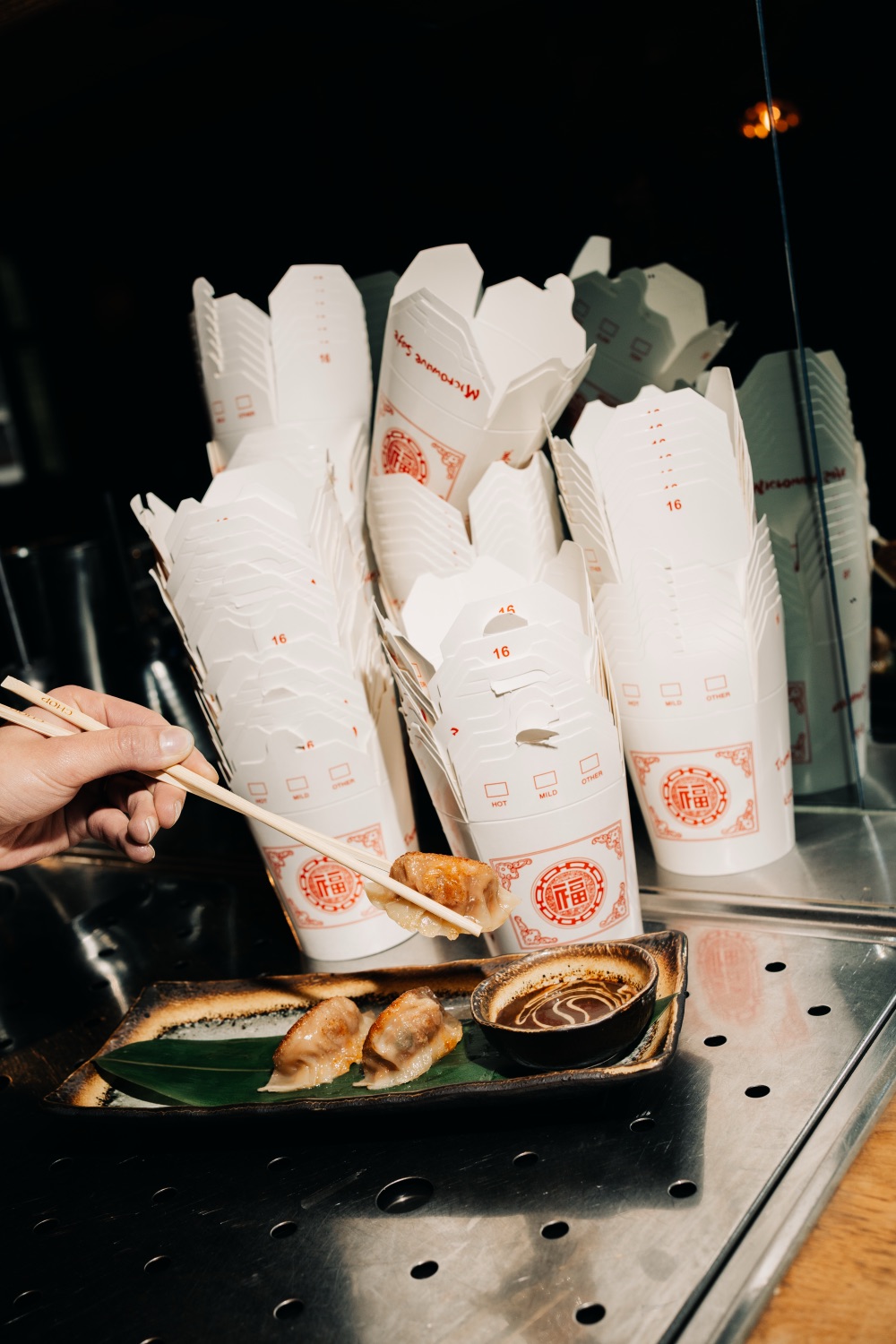 Chopsticks lifting a pan-fried dumpling next to a plate and stacks of takeout boxes