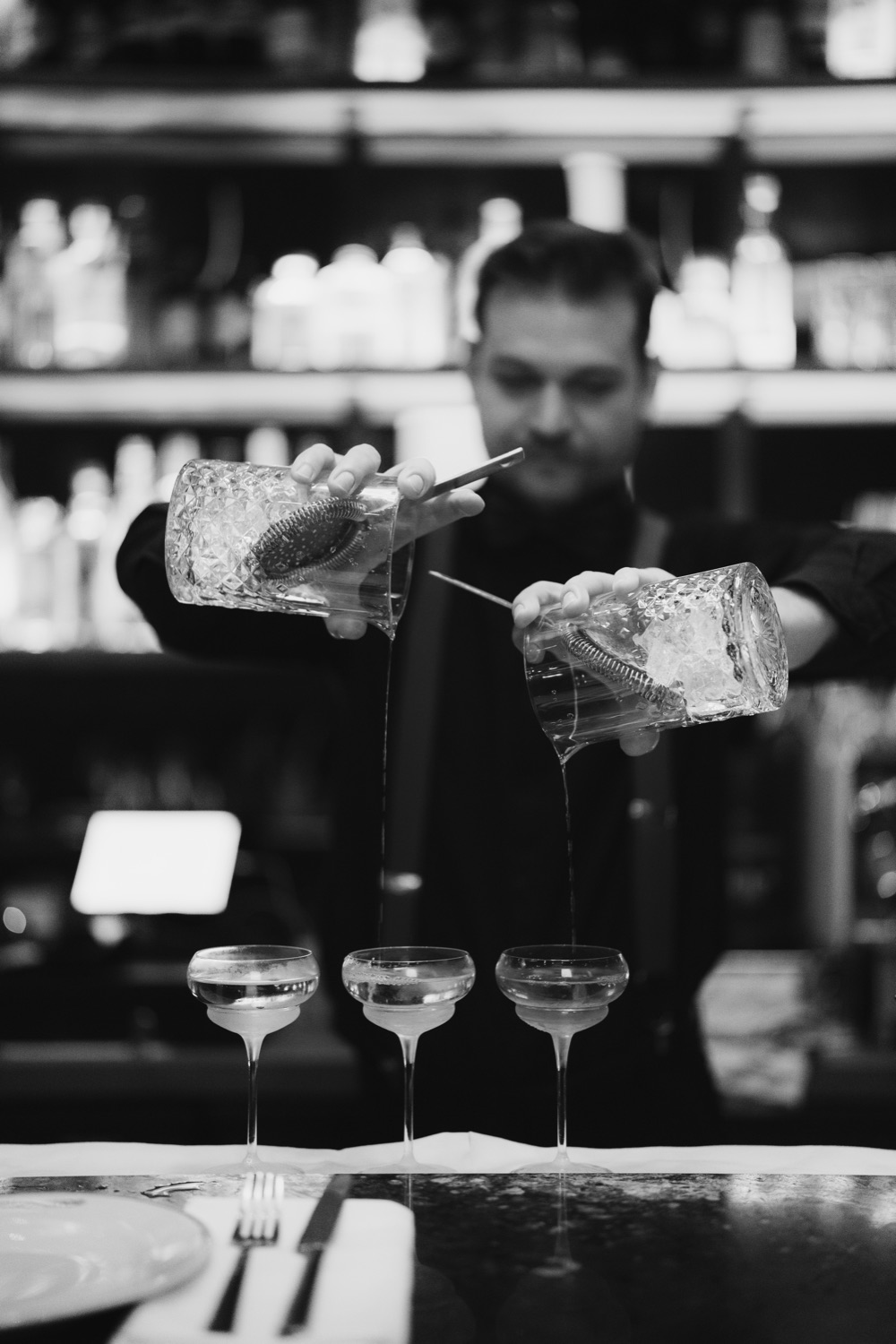 Zwart-witfoto van een bartender die cocktails inschenkt in drie coupe-glazen